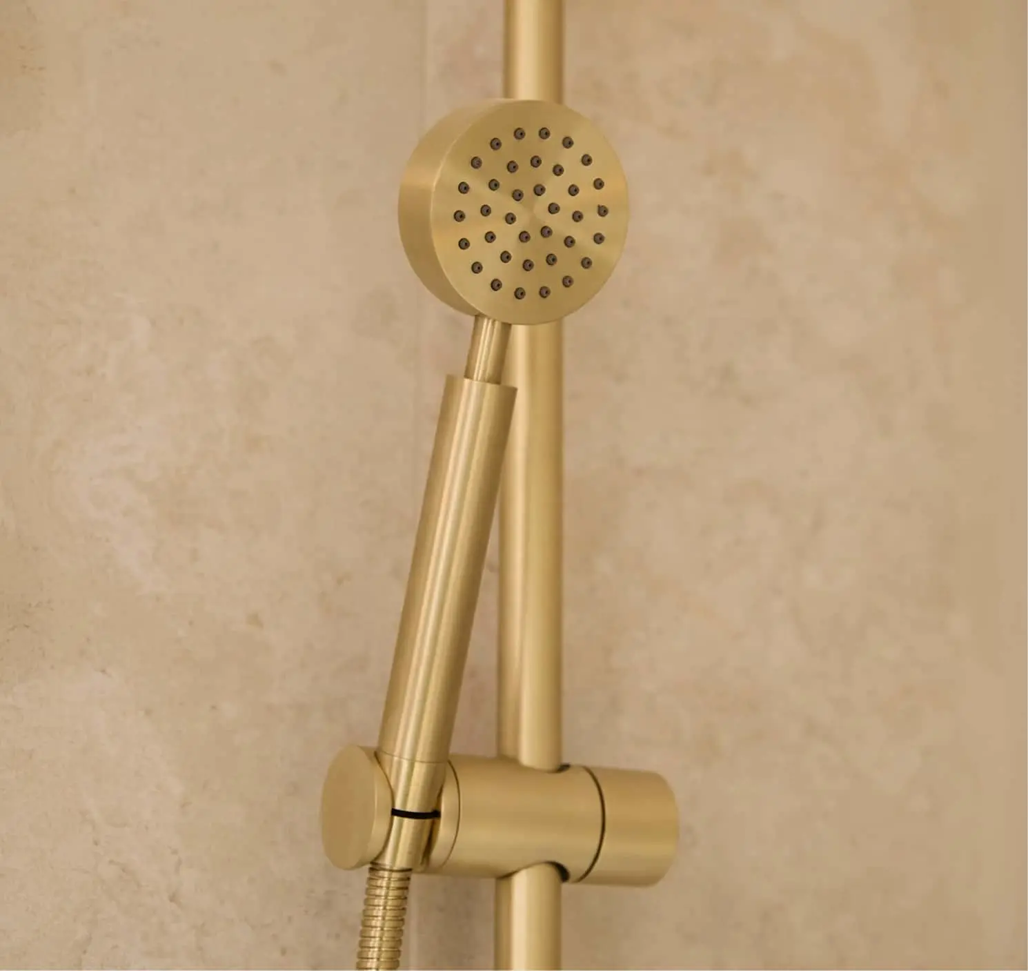Brushed gold handheld showerhead mounted on a vertical slide rail bar, against a beige stone wall.