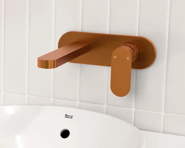 Copper-colored wall-mounted bathroom faucet with flat spout above a white Roca sink against white tiled wall.
