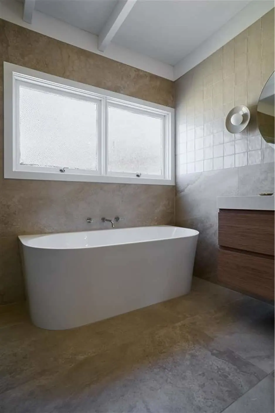 Modern bathroom with freestanding white bathtub, frosted windows, beige walls, white tile, and wooden vanity.