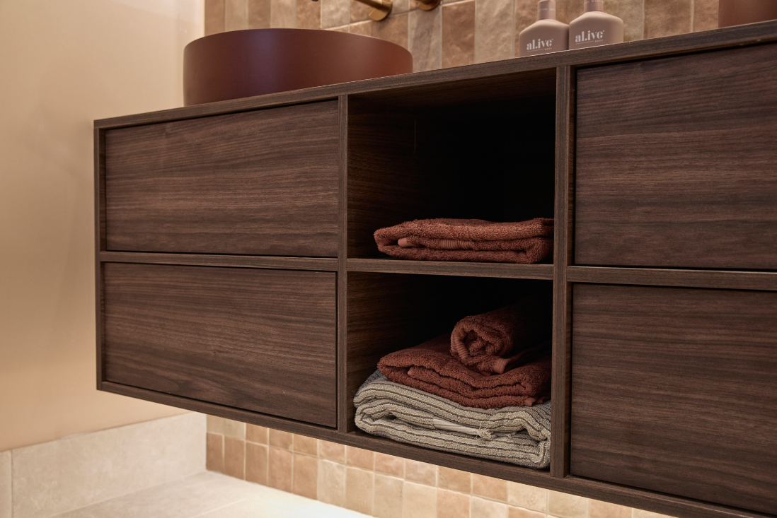 Dark wooden bathroom cabinet with folded brown and gray towels in open shelving and decorative bottles on top.