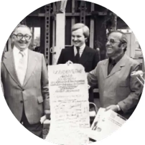 Three men in suits smiling while holding a document, standing outside a building in a black and white photograph.