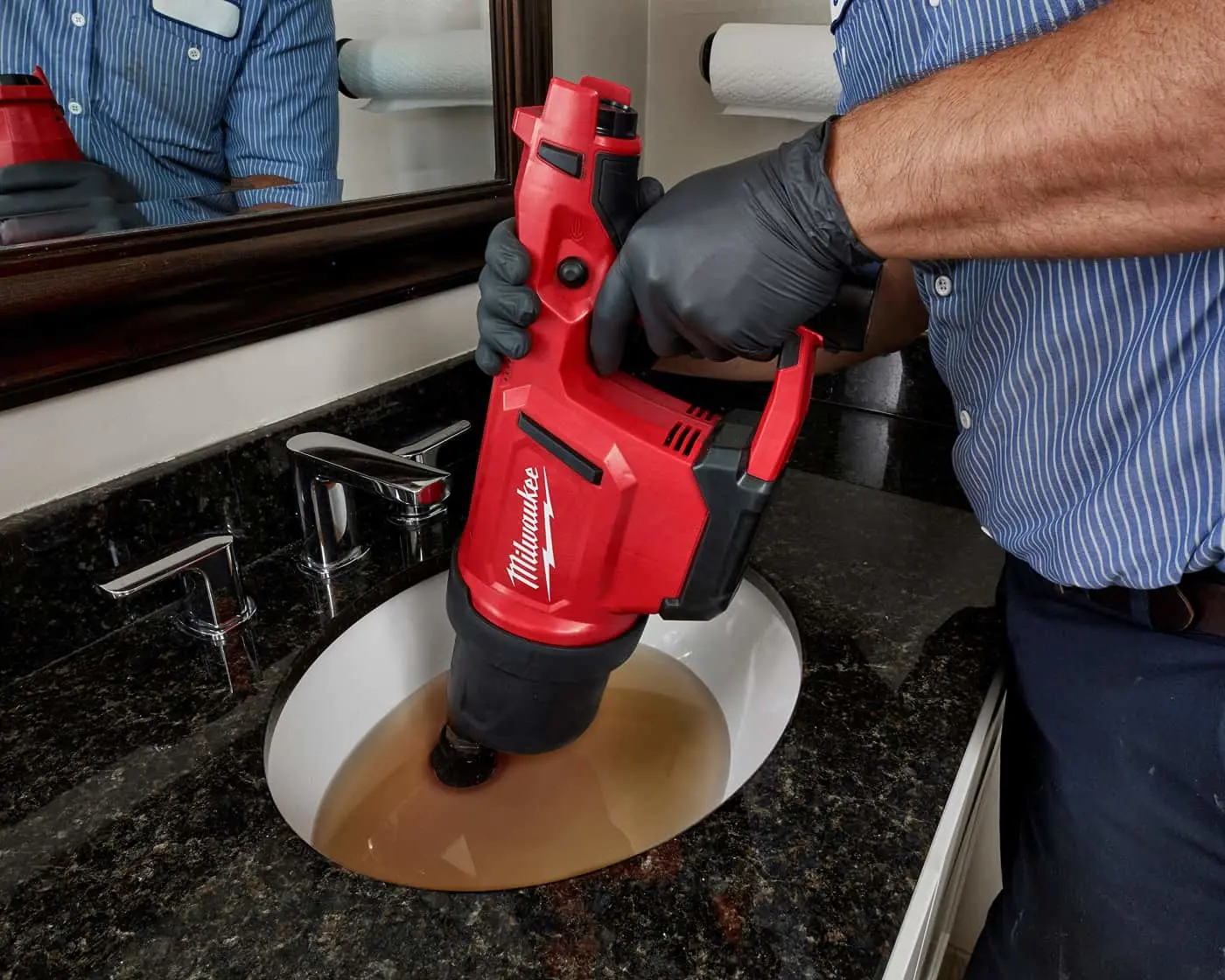Person in blue shirt using red Milwaukee drain cleaning tool on clogged bathroom sink with black gloves on.