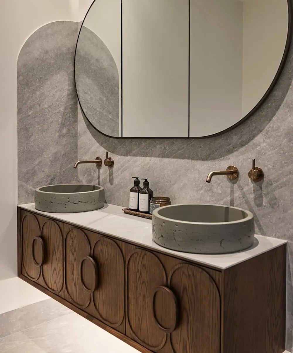 Modern bathroom with wooden vanity, concrete sinks, curved mirror, and copper wall-mounted faucets against gray stone wall.