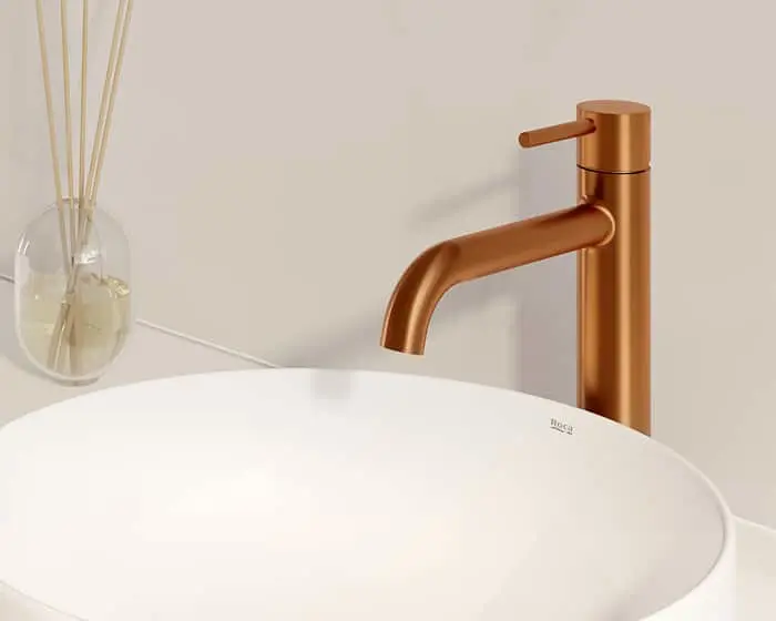 Bronze bathroom faucet with curved spout over a white basin sink, with reed diffuser visible in background.