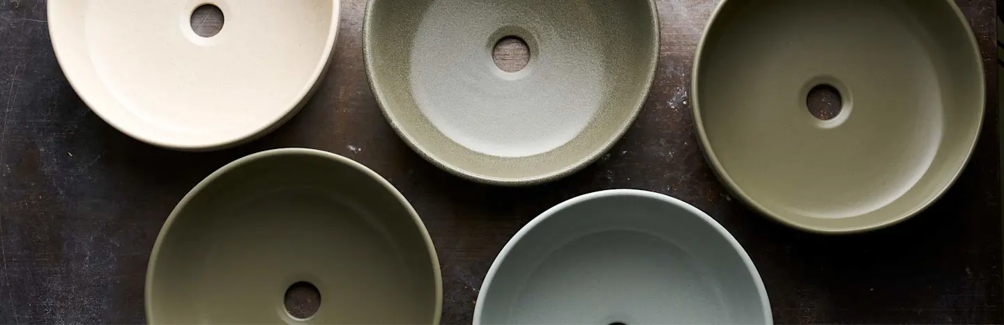 Five ceramic vessel sinks in various neutral tones arranged on a dark surface, viewed from above.