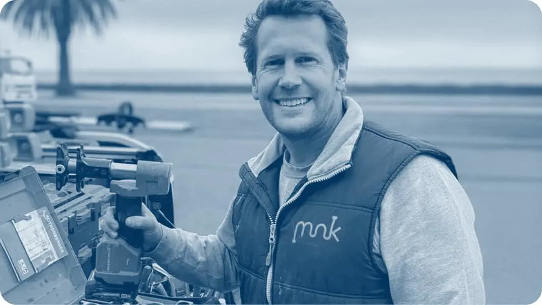 Person in blue-tinted photo wearing vest operating equipment near water with palm tree visible.