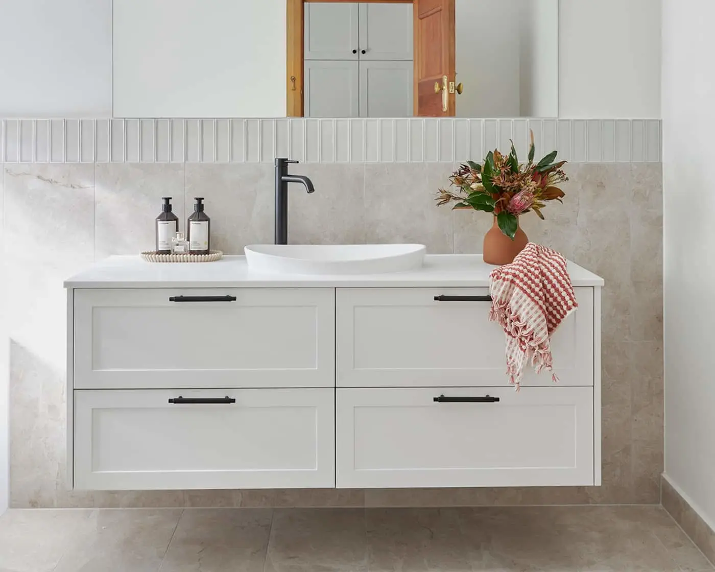 Modern white bathroom vanity with vessel sink, black faucet, terracotta vase with flowers, and red striped hand towel.