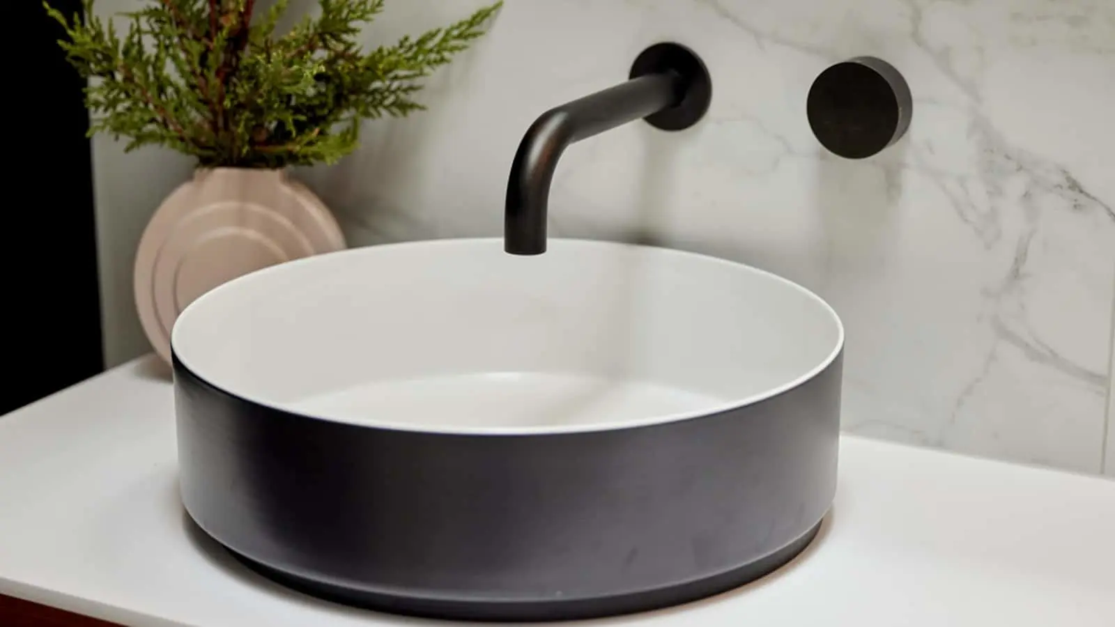 Modern circular sink with black faucet on white countertop, marble backsplash, and small plant in pink vase nearby.
