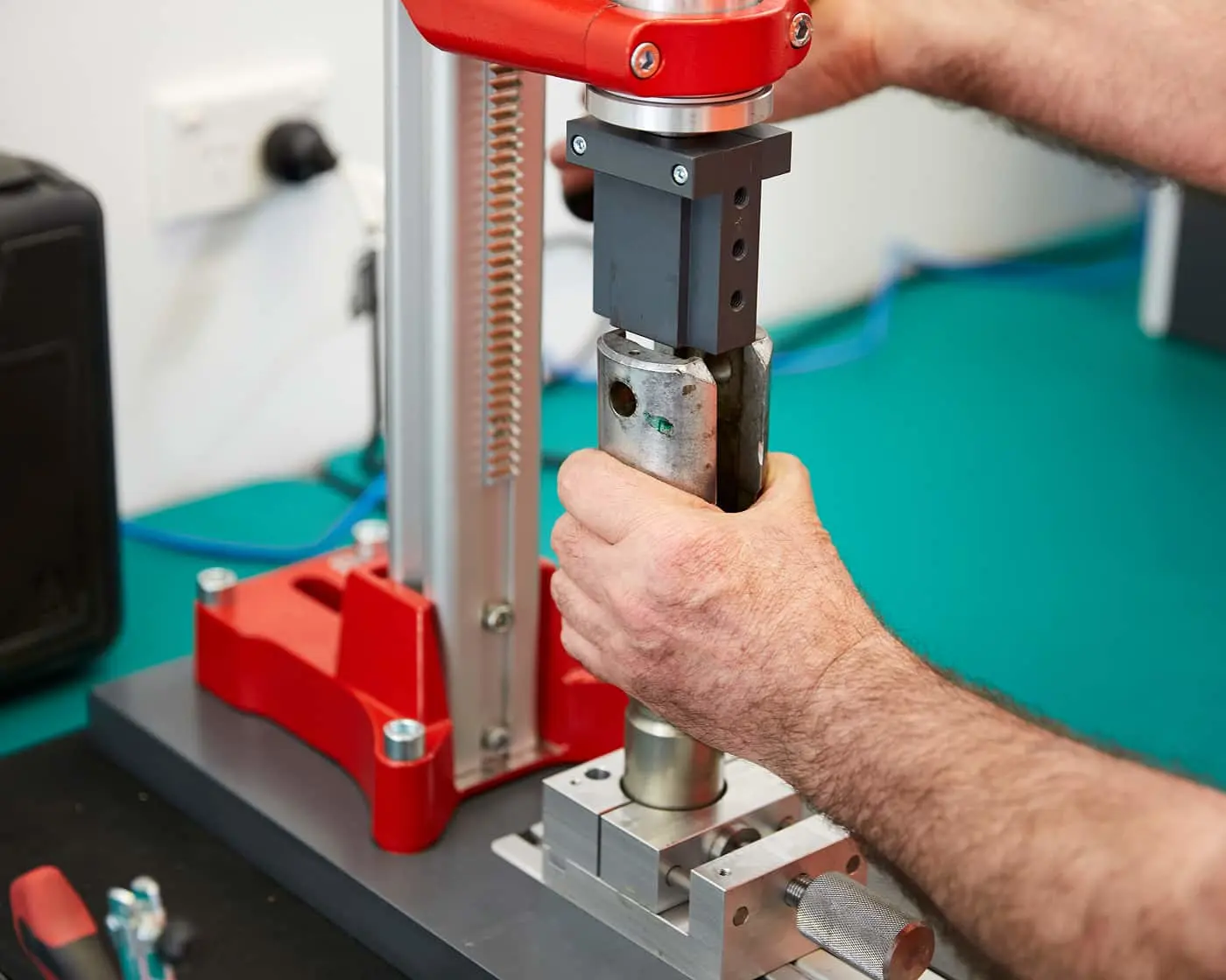 Hands operating a material testing machine with red and silver components on a turquoise surface in a laboratory setting.
