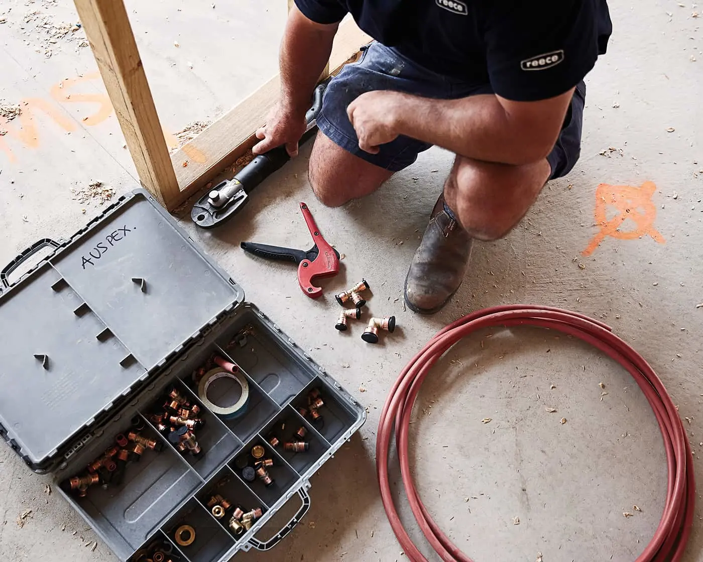 Construction worker installing plumbing, with toolbox of copper fittings, pipe cutter, and red tubing on concrete floor.