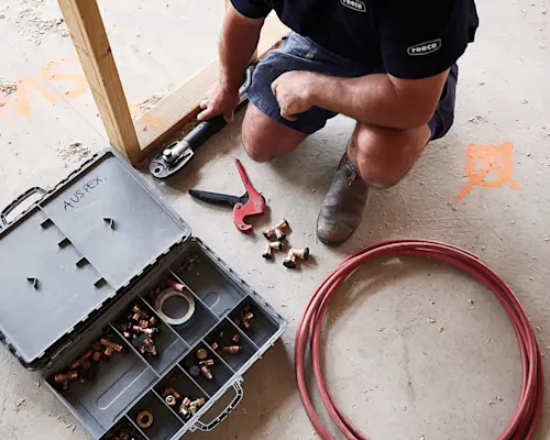 Construction worker installing plumbing, with toolbox of copper fittings, pipe cutter, and red tubing on concrete floor.