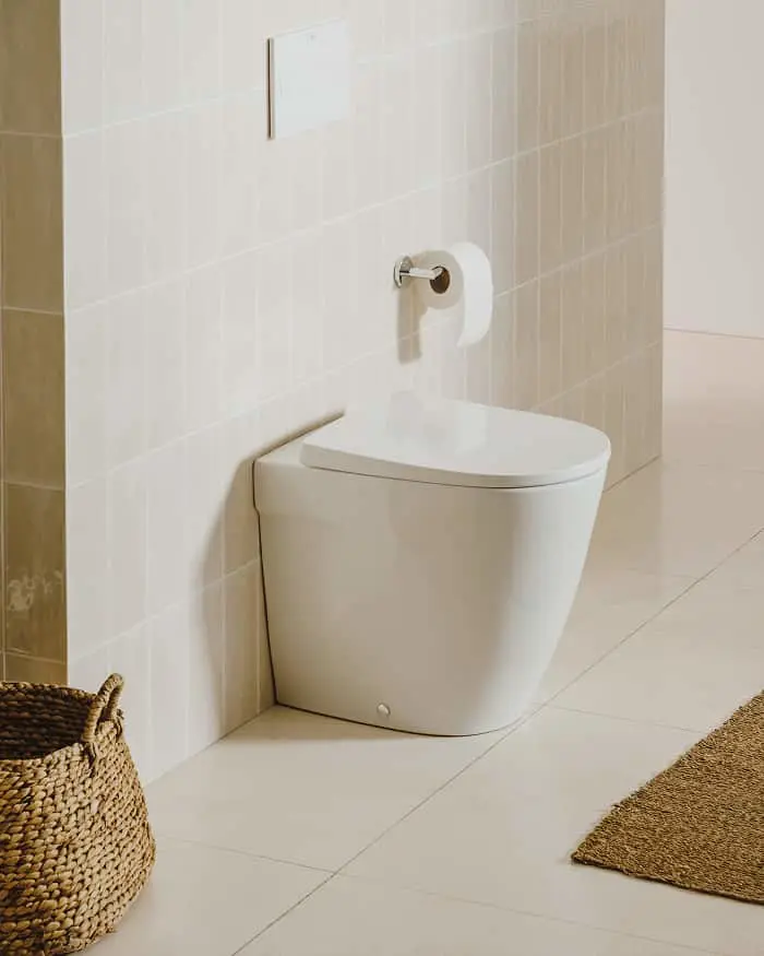 Modern white toilet with toilet paper holder on beige tiled wall, woven basket and jute mat visible.