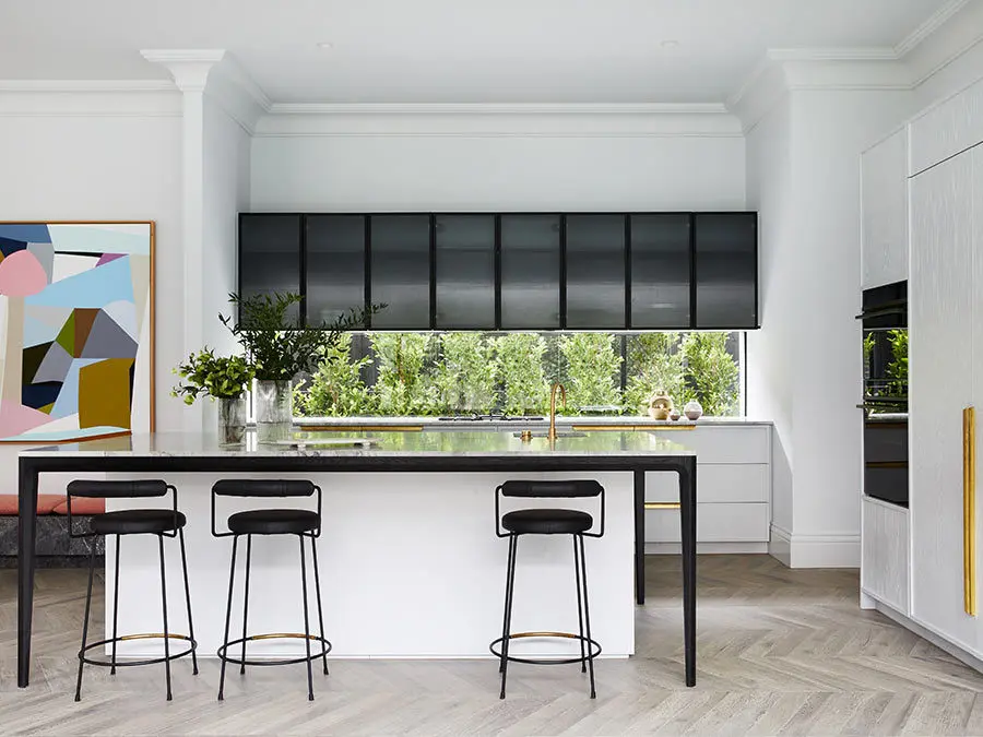 Modern white kitchen with black bar stools, window overlooking greenery, colorful abstract art, and herringbone flooring.