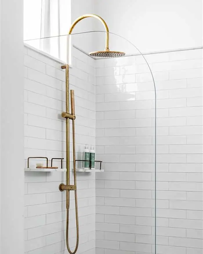 Modern white tiled shower with gold rainfall showerhead, handheld attachment, glass partition, and built-in shelving.