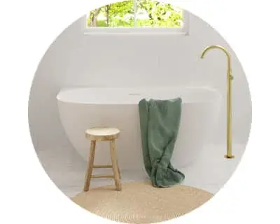 Minimalist white bathroom with freestanding tub, wooden stool, green towel, and brass fixtures under a window.