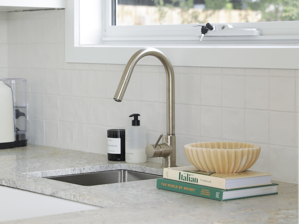 Modern kitchen sink with brushed nickel faucet, marble countertop, cookbooks, and a decorative bowl beneath a window.