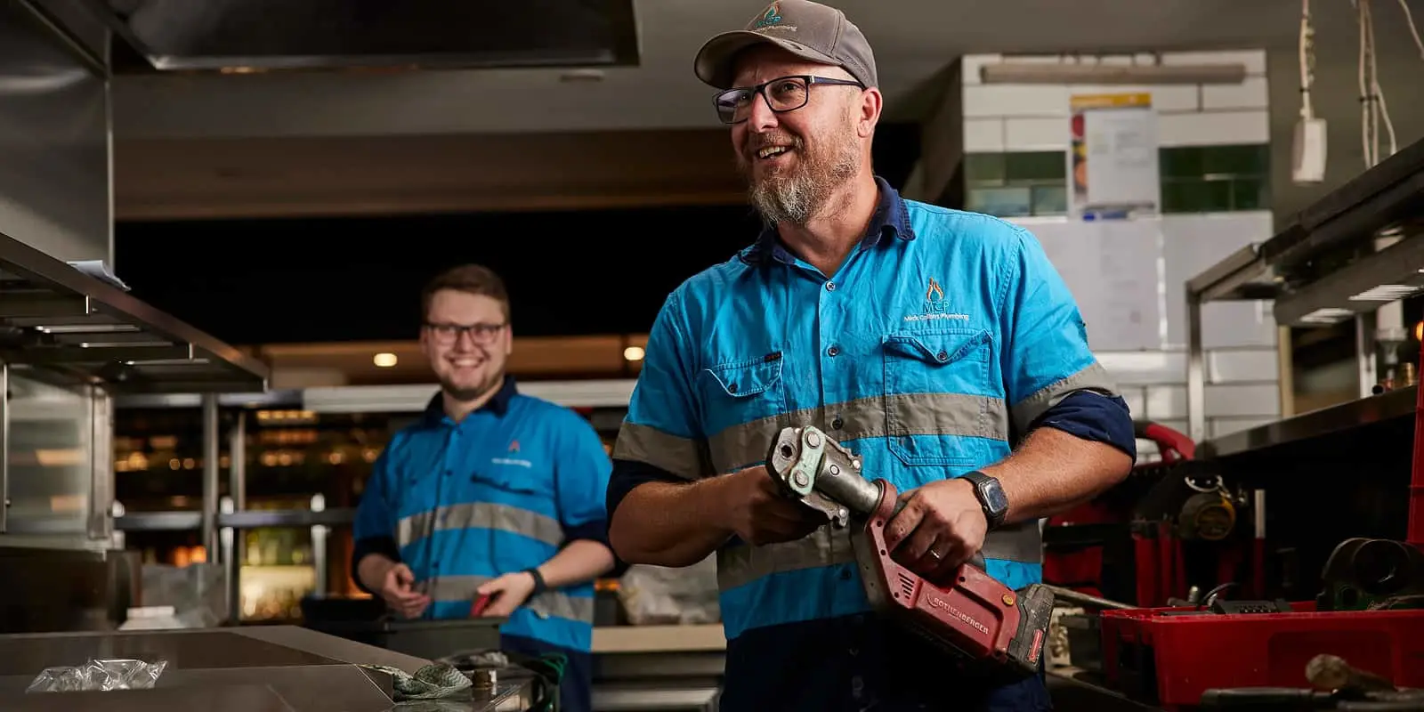 Two workers in blue uniforms with reflective stripes in a workshop, one holding a power tool, both smiling.