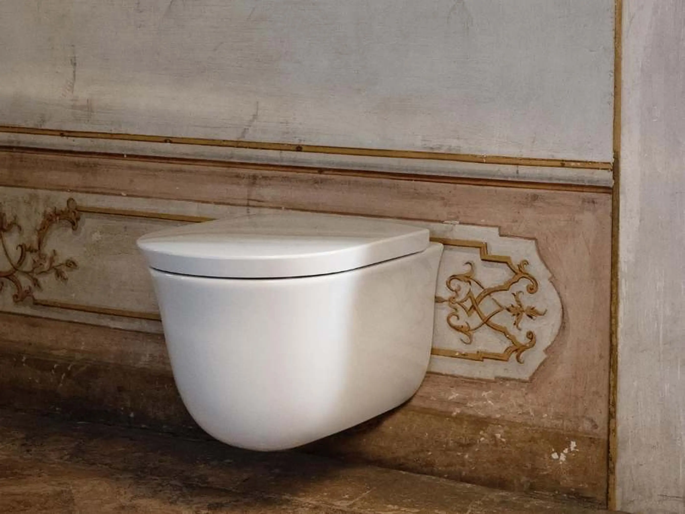 Modern white wall-mounted toilet against ornate beige wall with gold decorative accents in an elegant bathroom.