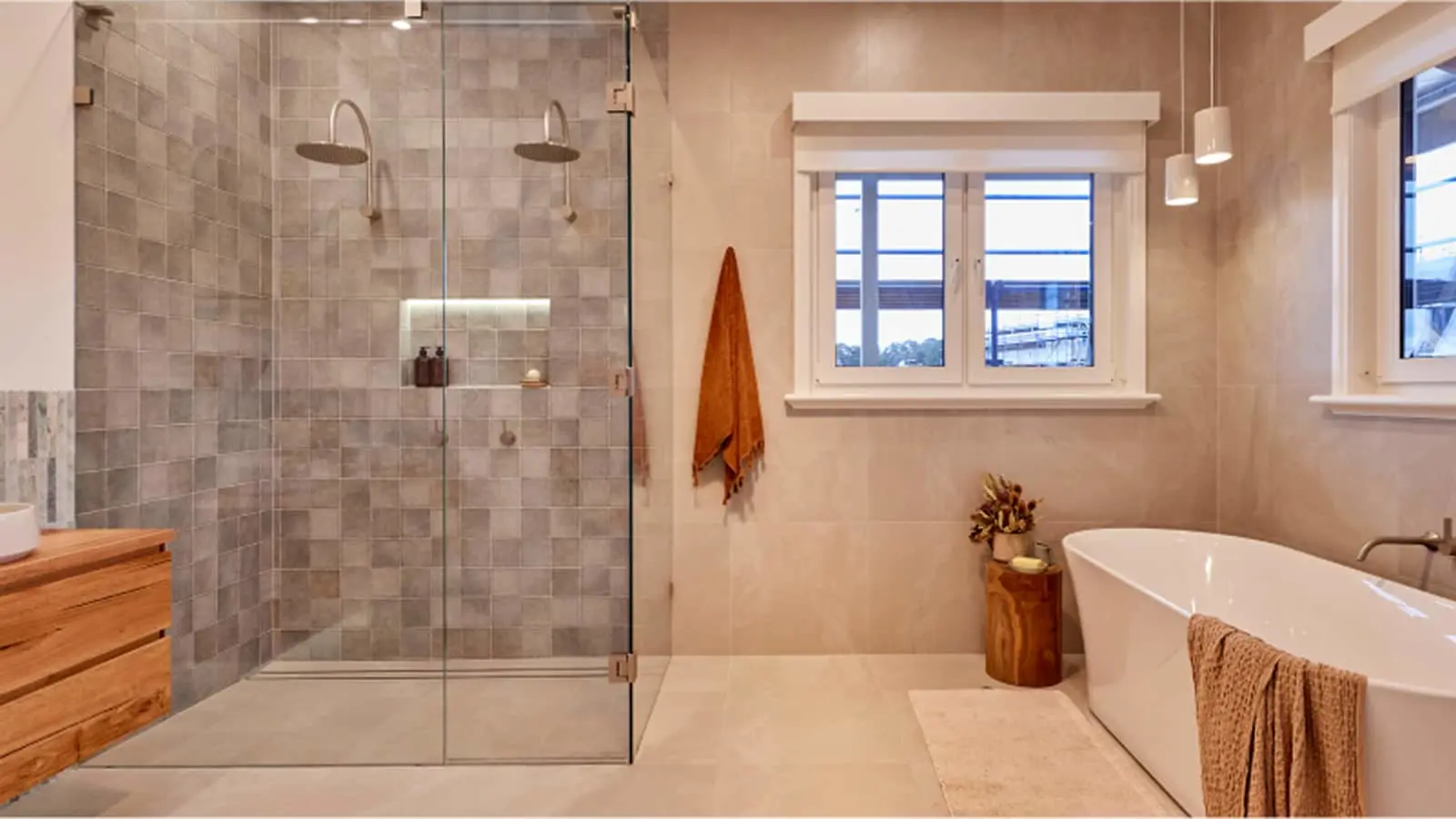 Modern bathroom with glass shower, freestanding white tub, gray tile walls, wooden accents, and natural light from windows.