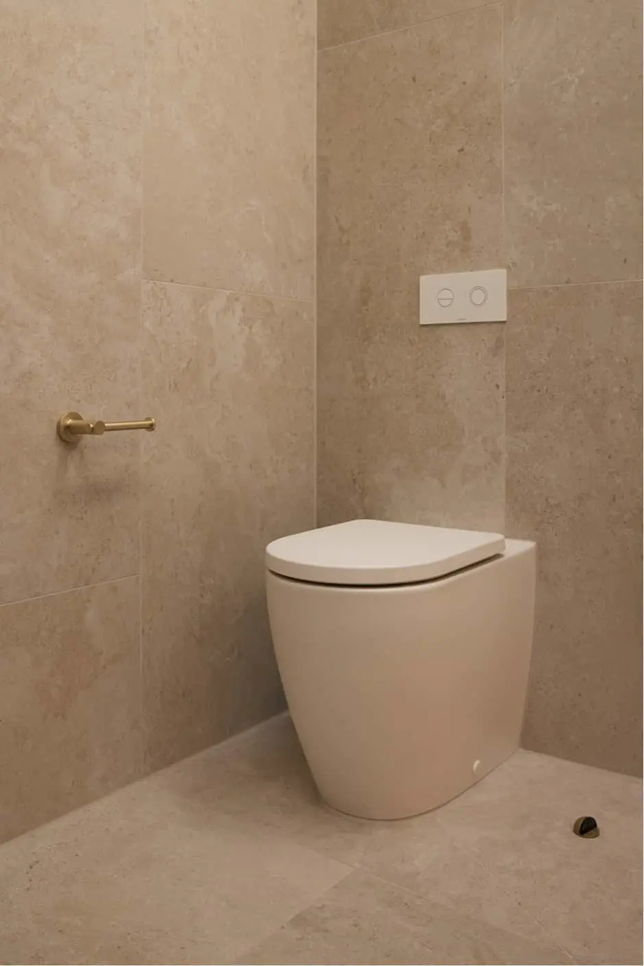 White toilet in corner with beige tile walls, brass toilet paper holder, and white dual flush button panel.