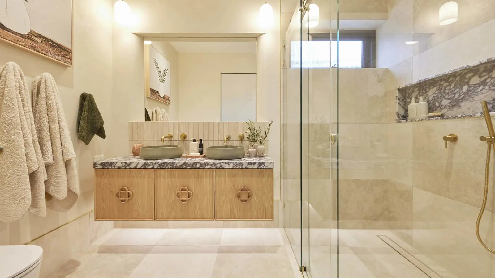 Modern bathroom with wooden vanity, dual vessel sinks, marble countertop, glass shower enclosure, and beige tile floors.