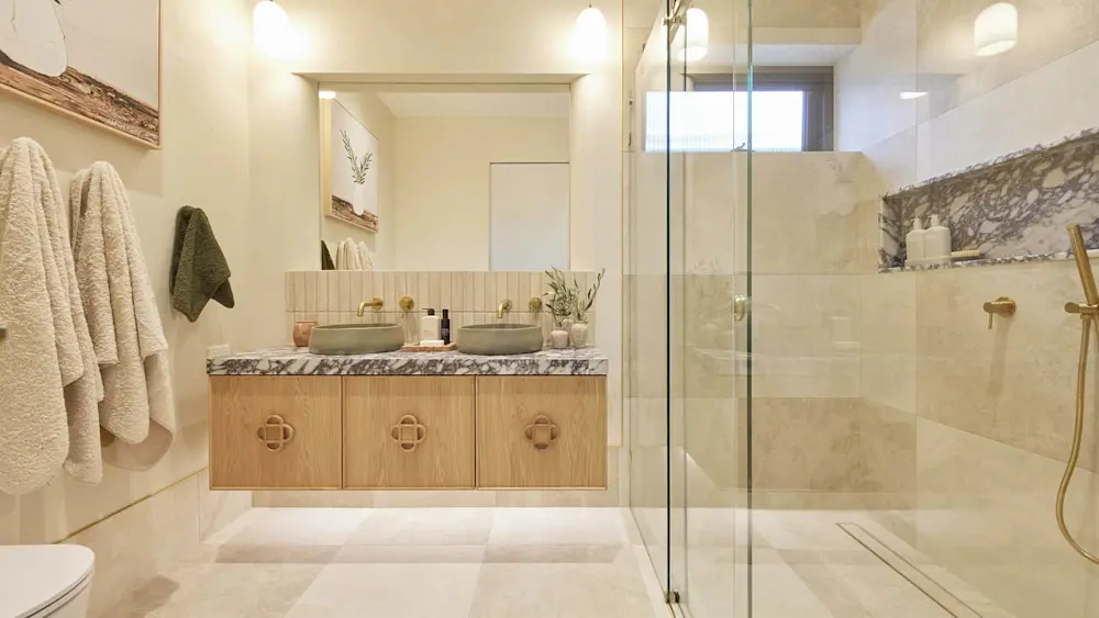 Modern bathroom with wooden vanity, dual vessel sinks, marble countertop, glass shower enclosure, and beige tile floors.