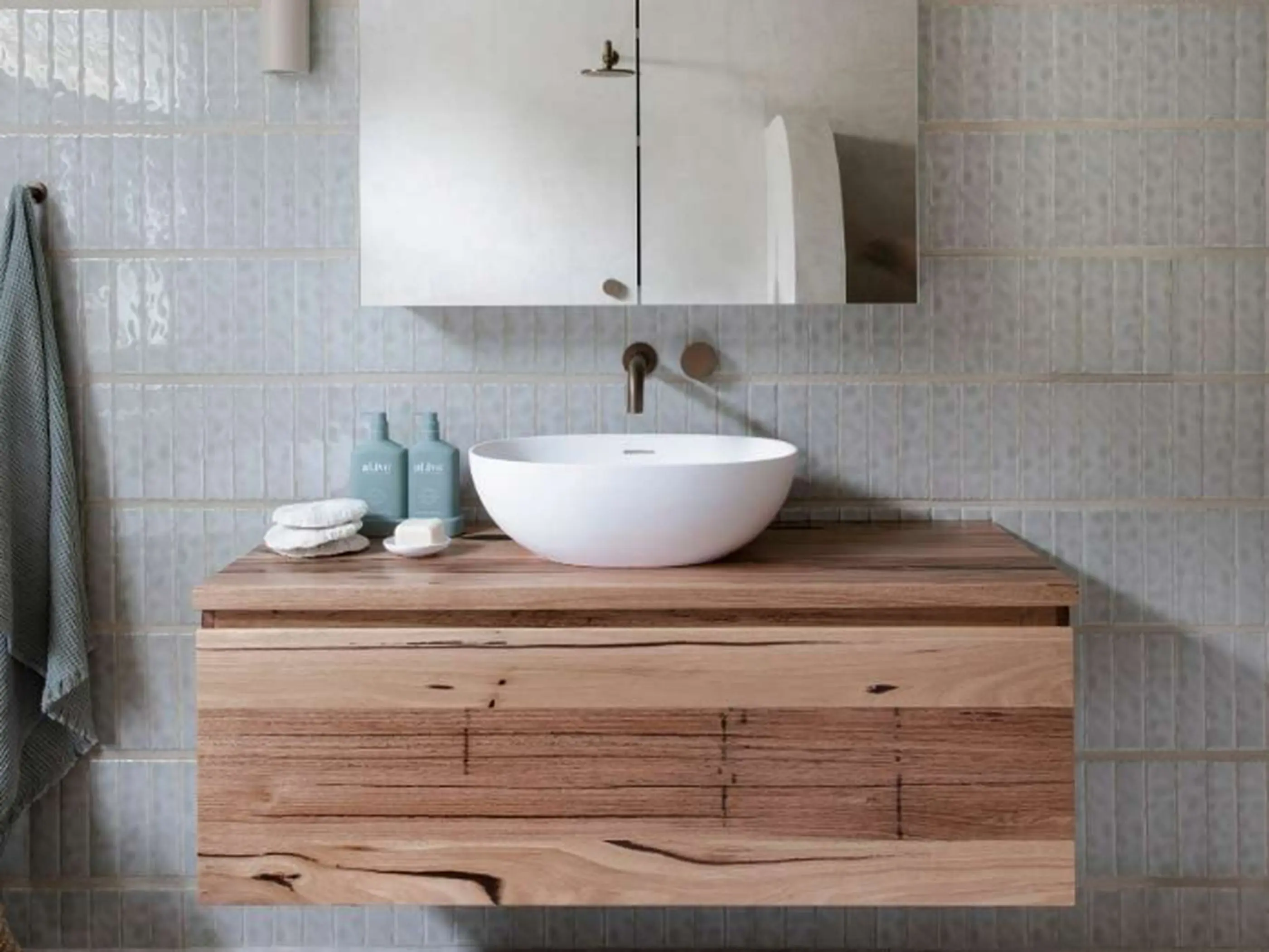 Modern bathroom with white vessel sink on wooden vanity, pale blue tiled walls, and wall-mounted brass faucet.