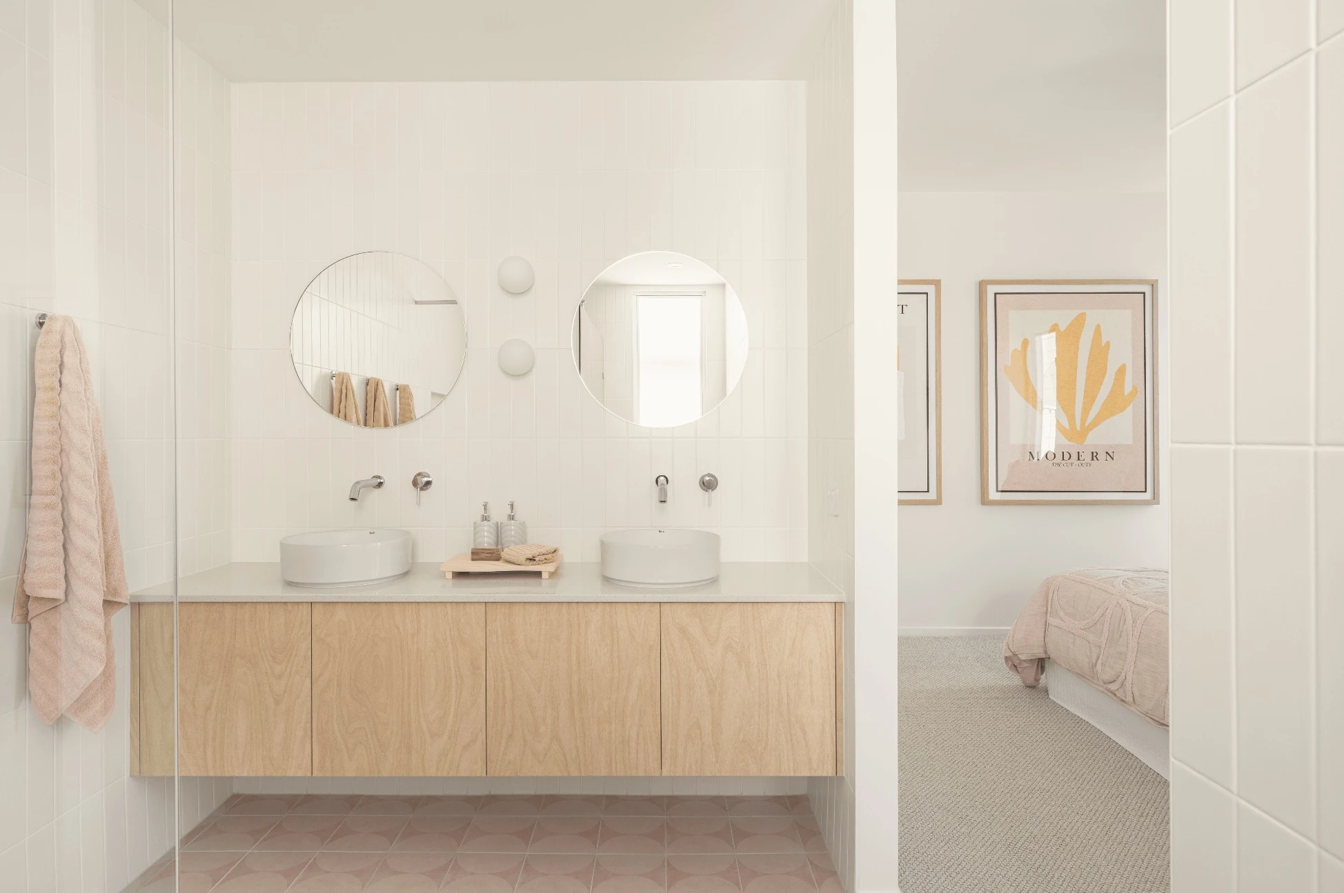 Modern bathroom with dual sinks, round mirrors, wooden vanity, and view into bedroom with abstract wall art.