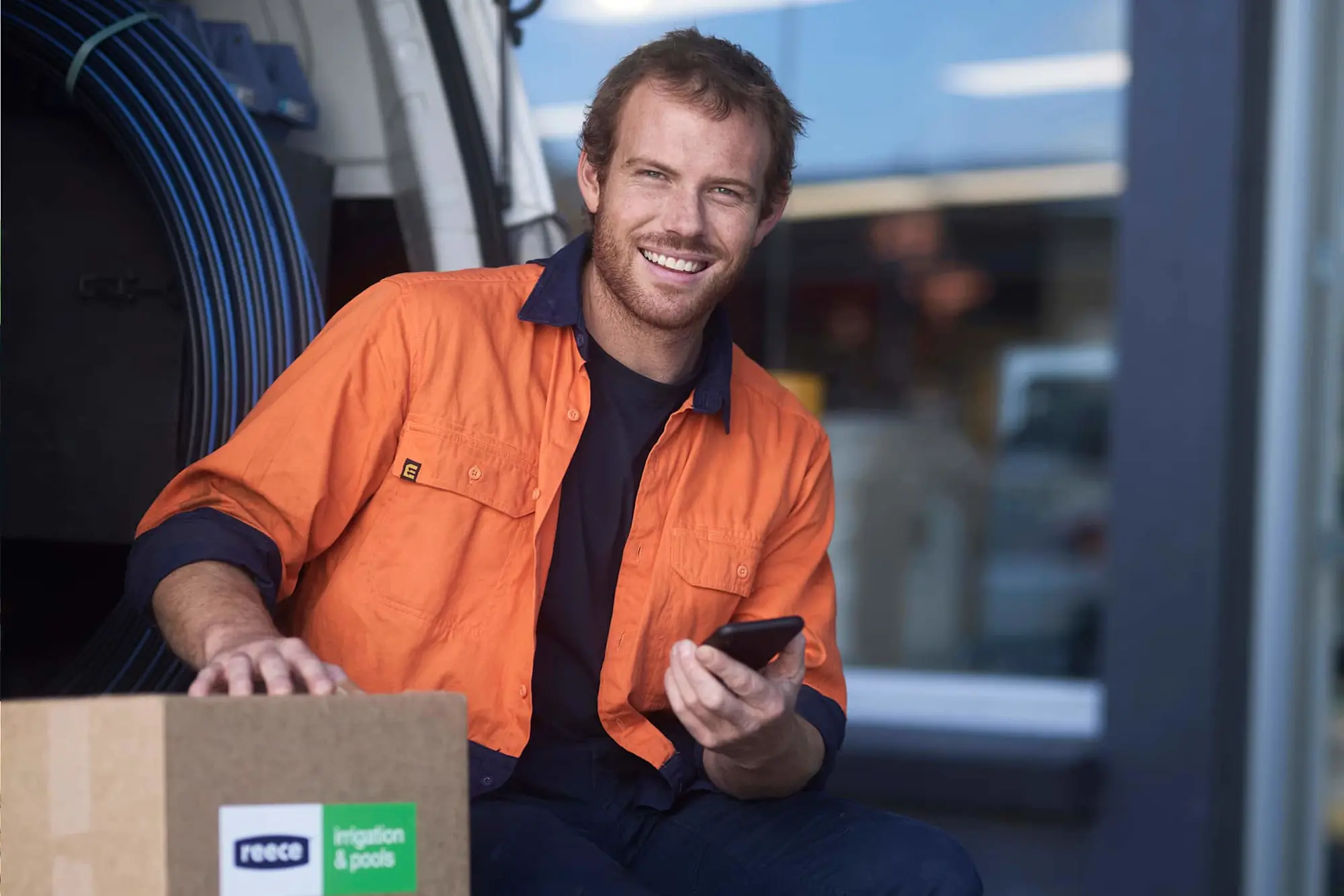 Person in bright orange work shirt smiling while holding a phone, with blue cables and a cardboard box nearby.