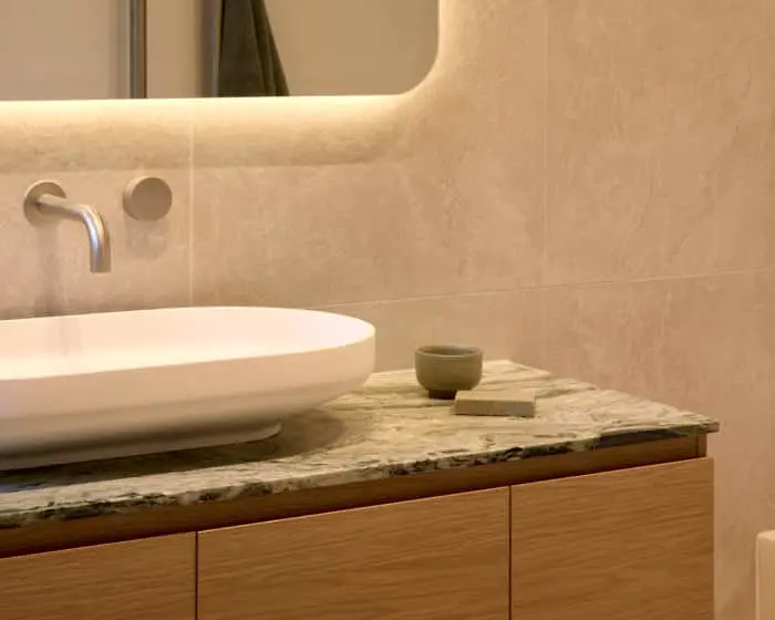 Modern bathroom with white vessel sink on stone countertop, wall-mounted faucet, and backlit mirror above beige tiles.