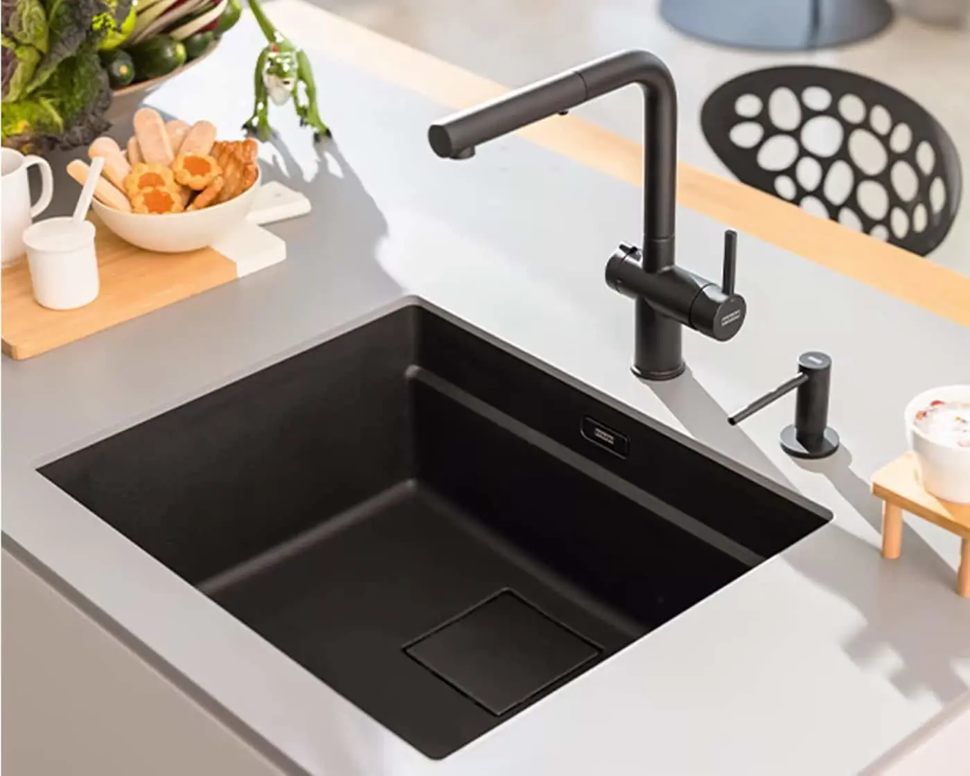 Modern black kitchen sink with matching faucet on white countertop, bowl of cookies nearby and fresh vegetables visible.