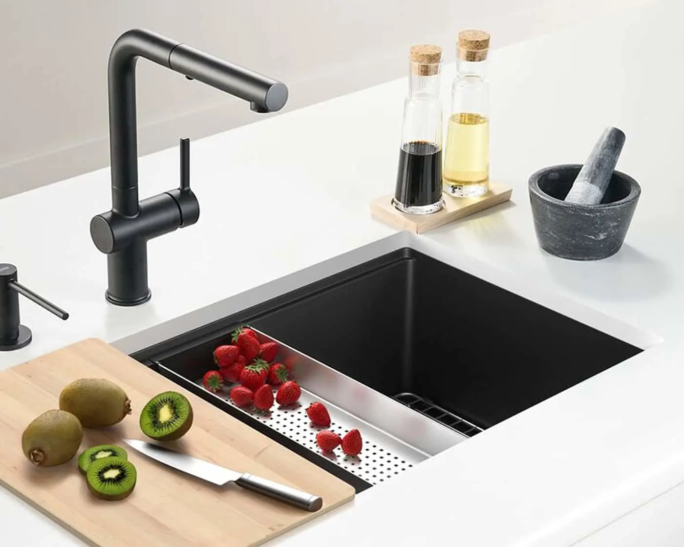 Modern black kitchen sink with matte faucet, strawberries in colander, kiwi on cutting board, and oil bottles nearby.