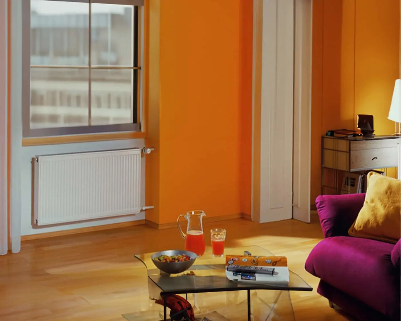 Modern living room with orange walls, purple sofa, glass coffee table, and window with radiator beneath.