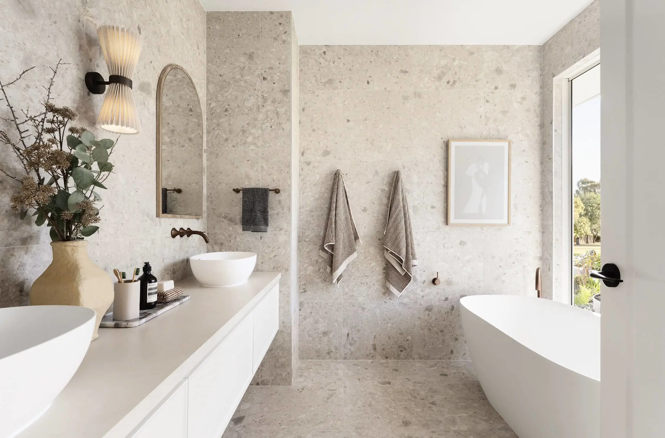 Modern bathroom with stone walls, white vanity, freestanding tub, wall sconce, and hanging towels beside a window.