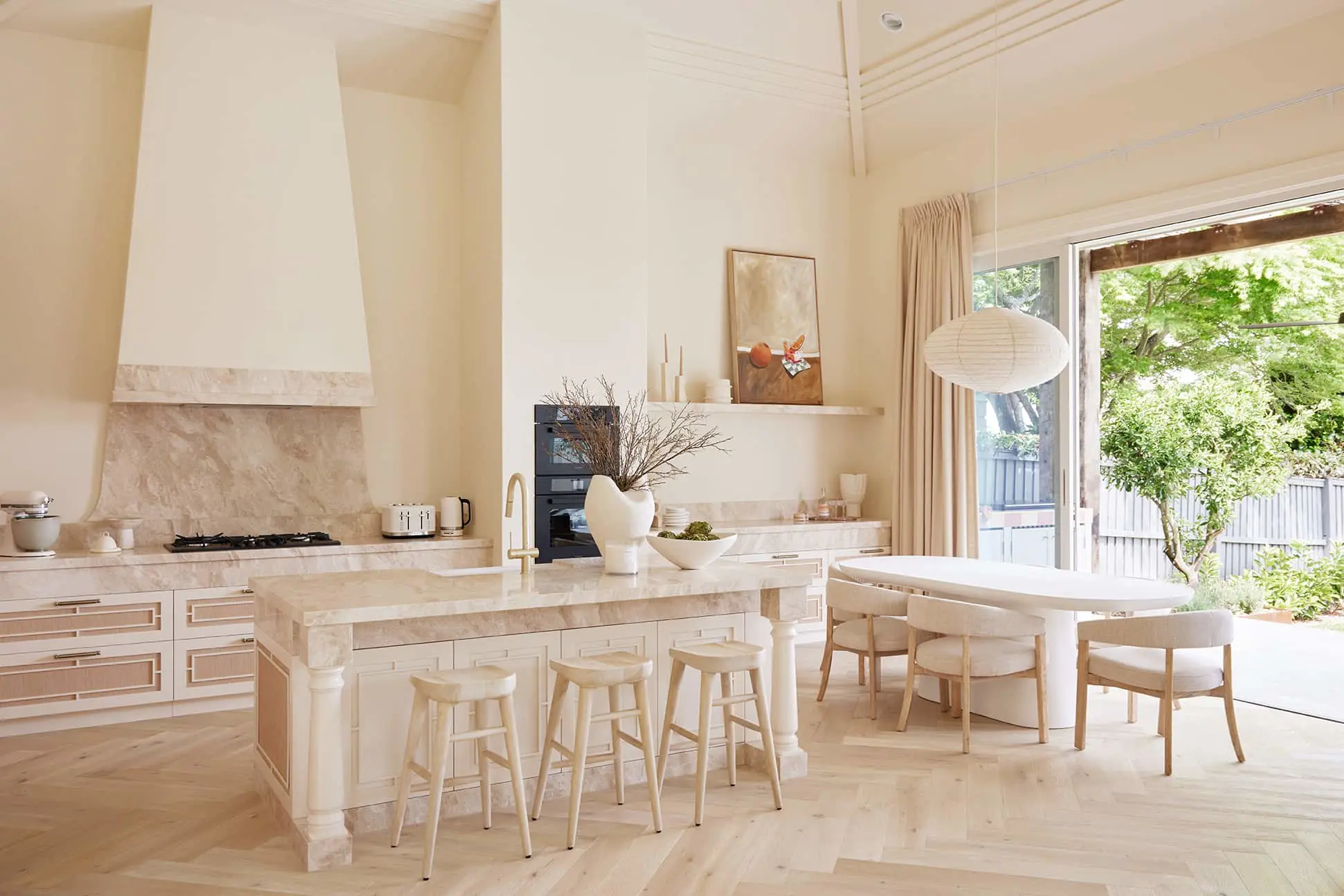 Modern cream-coloured kitchen with marble island, wooden stools, dining table, and large windows overlooking garden.