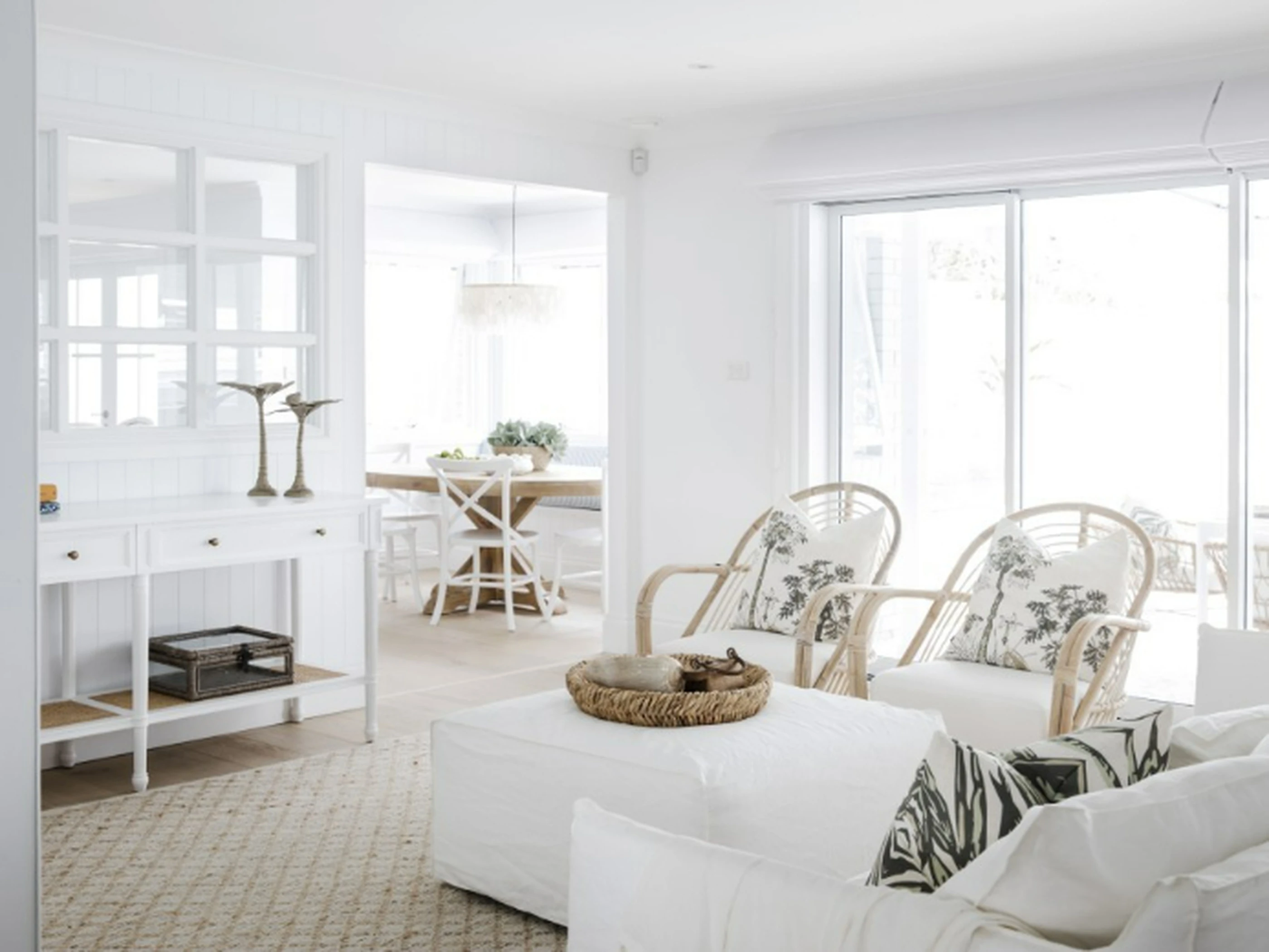 Bright white coastal living room with rattan chairs, white ottoman, and dining area visible through doorway with large windows.