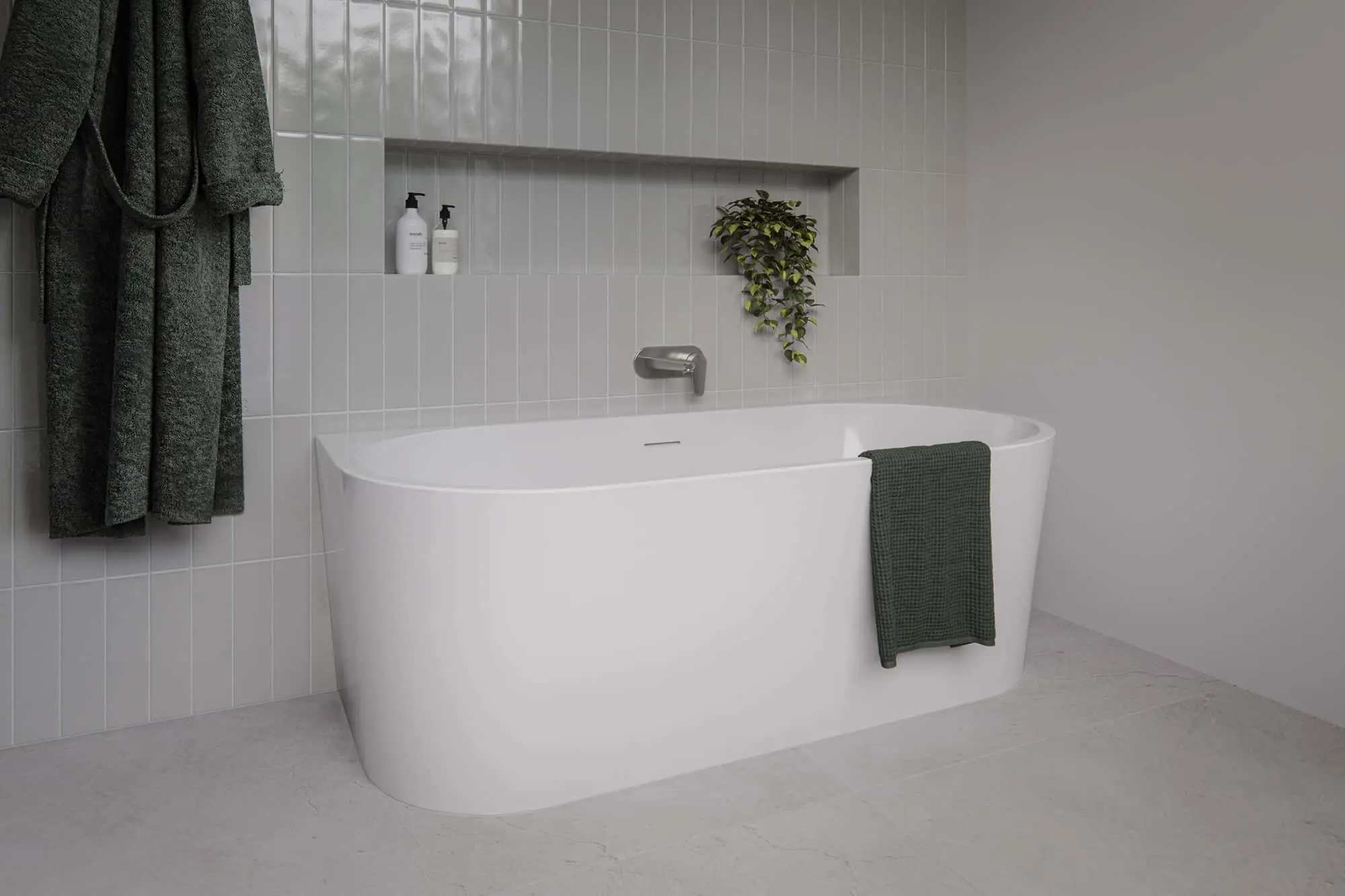 White freestanding bathtub with green towel against white tiled wall with recessed shelf holding bottles and plant.