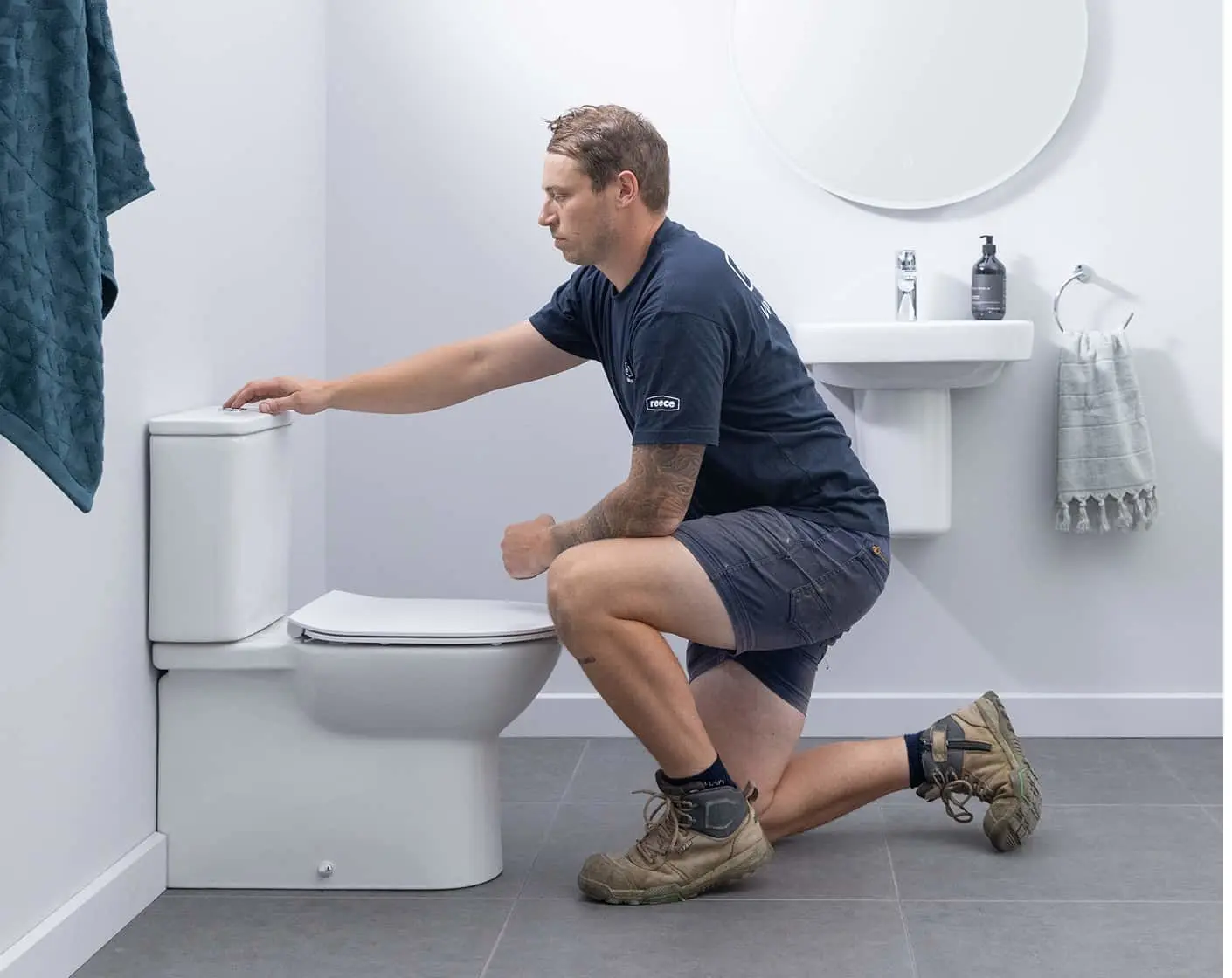 Plumber in navy uniform kneeling beside toilet in modern white bathroom with sink and blue towel visible.