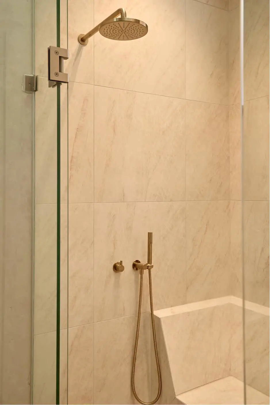 Modern shower with beige marble tiles, brass rainfall showerhead, handheld sprayer, and glass door.