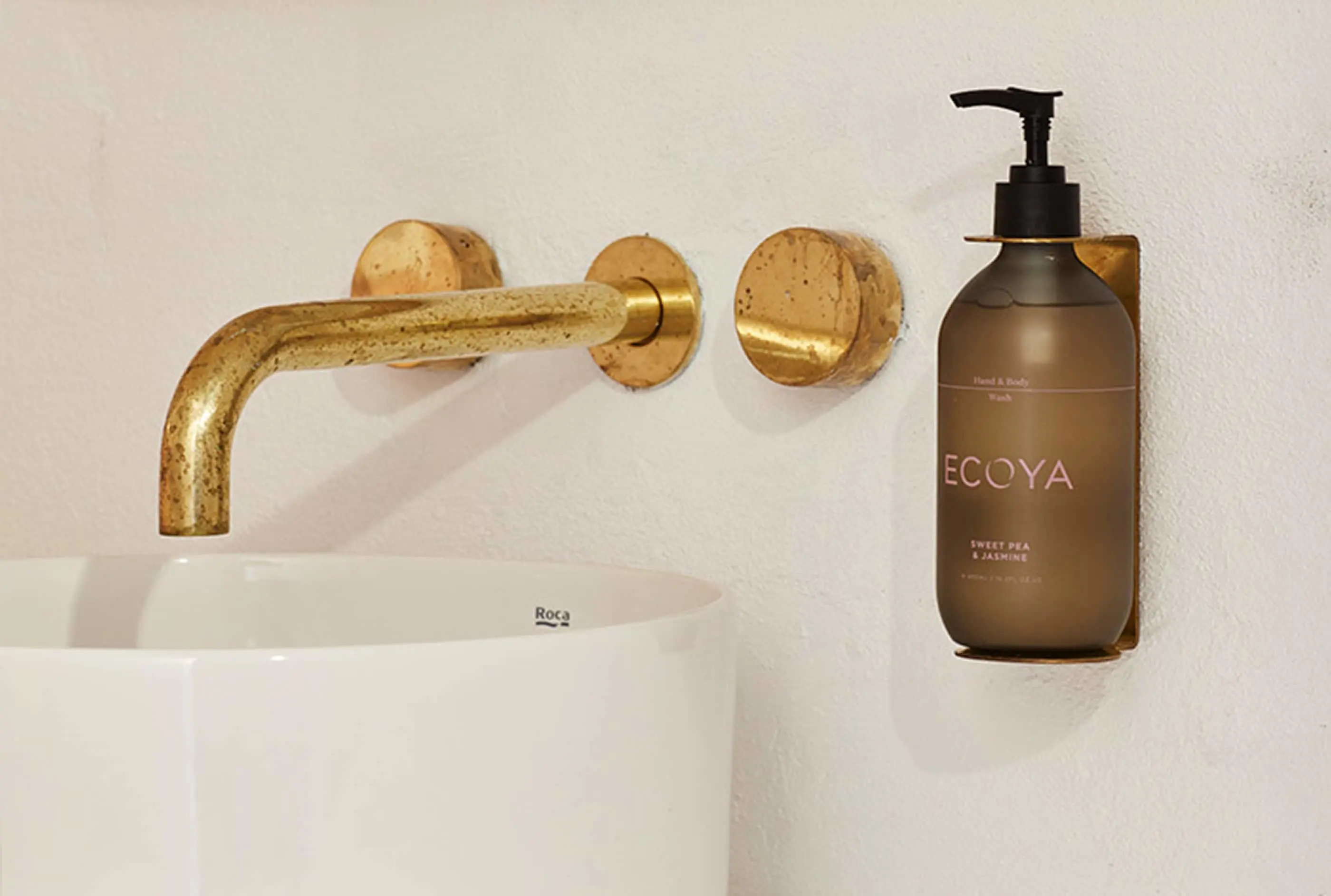 Bathroom sink with brass wall-mounted faucet and ECOYA hand soap dispenser in brown bottle mounted on white wall.
