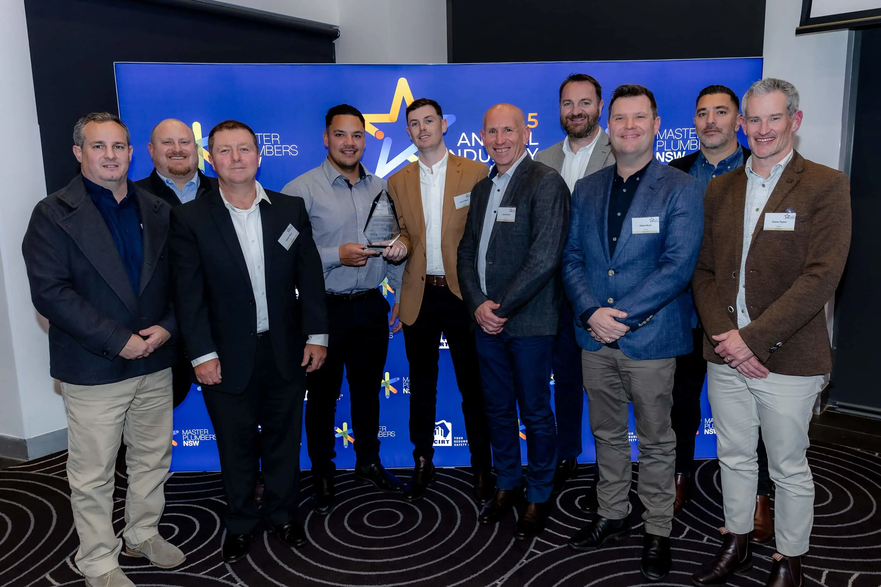 Group of men in business attire at Master Plumbers NSW industry awards event, one holding a trophy.