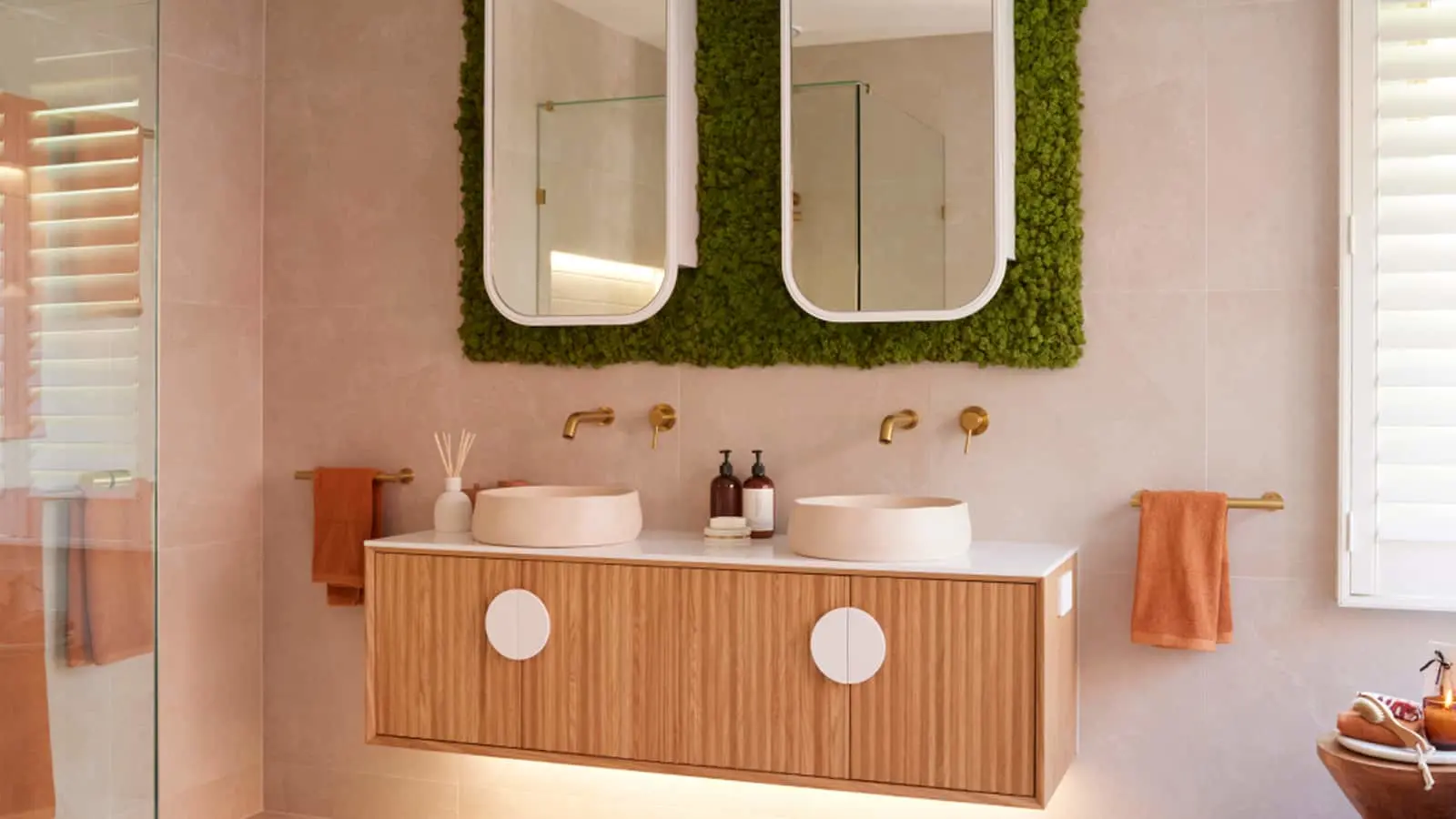 Modern bathroom with dual sinks, wooden vanity, moss wall behind mirrors, and gold fixtures with coral towels.