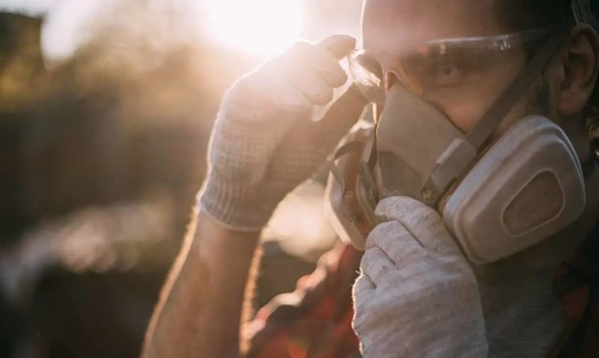 Person wearing safety goggles and respirator mask adjusting protective equipment in golden sunlight.