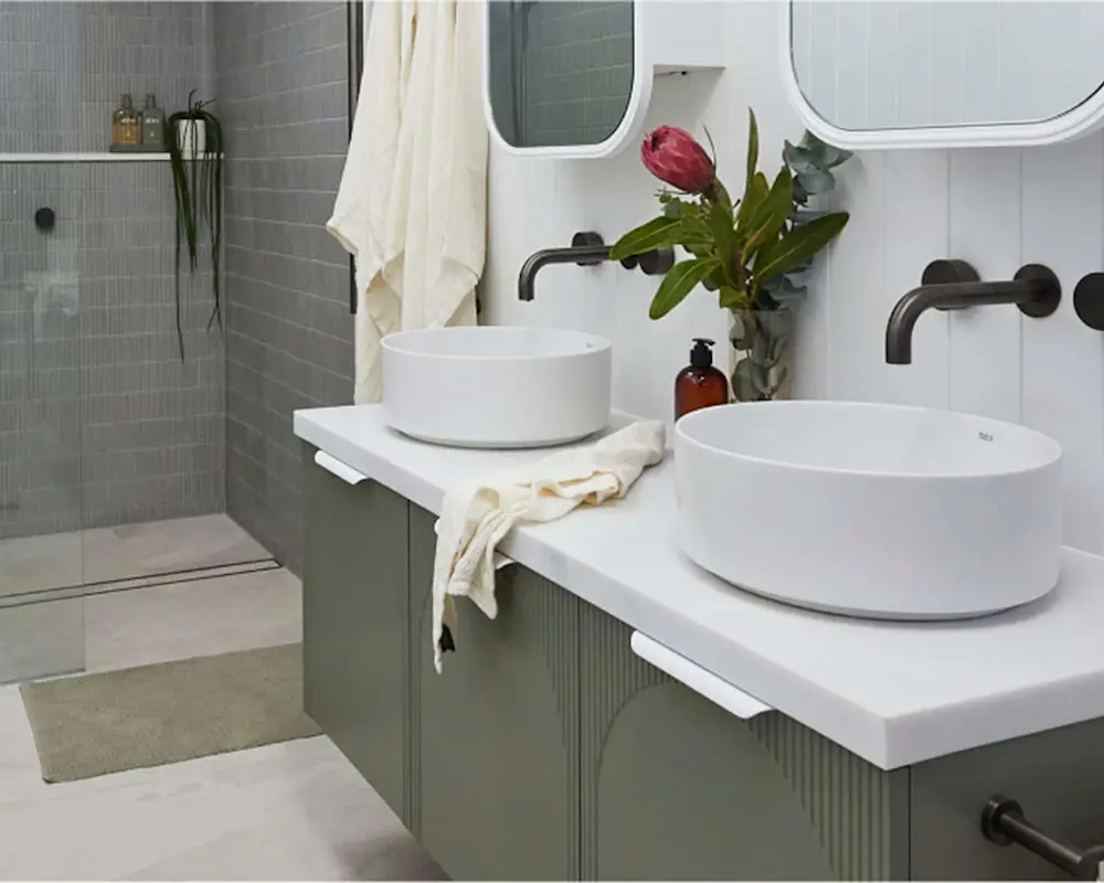Modern bathroom with dual white vessel sinks, gray tile walls, black fixtures, and a small vase with red flower.