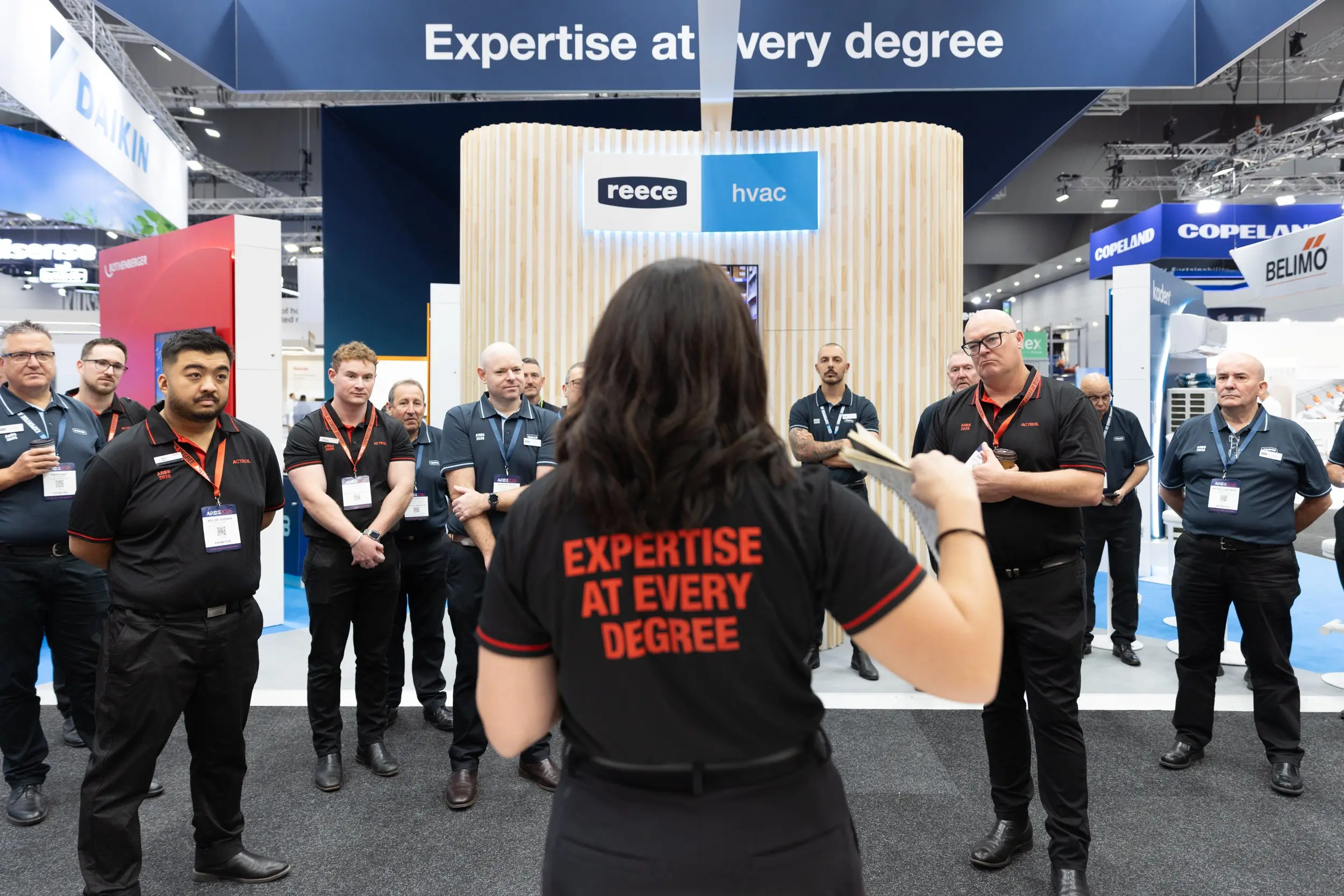 A woman addresses a group of staff at the Reece HVAC trade show booth, wearing a black shirt reading "Expertise at Every Degree."