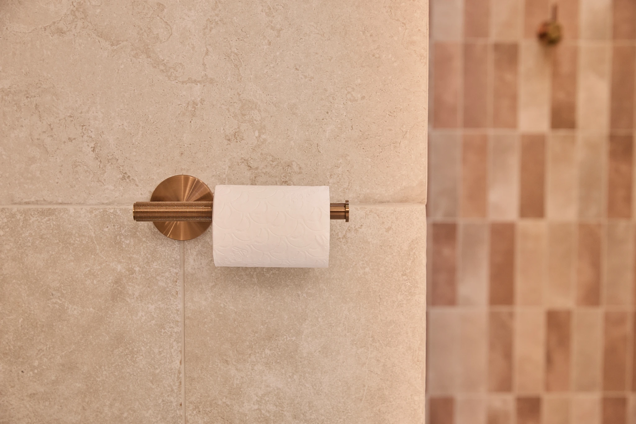Copper toilet paper holder mounted on beige stone bathroom wall with tiled wall visible in background.