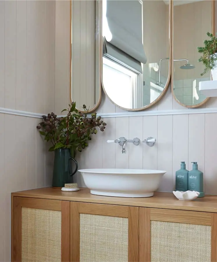 Modern bathroom with white vessel sink on wooden vanity, oval mirrors, green pitcher with plants, and teal soap bottles.