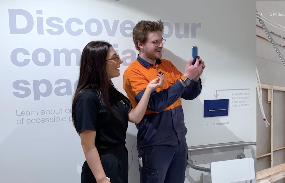 Two people looking at a phone in an accessible bathroom display area with informational wall text visible.