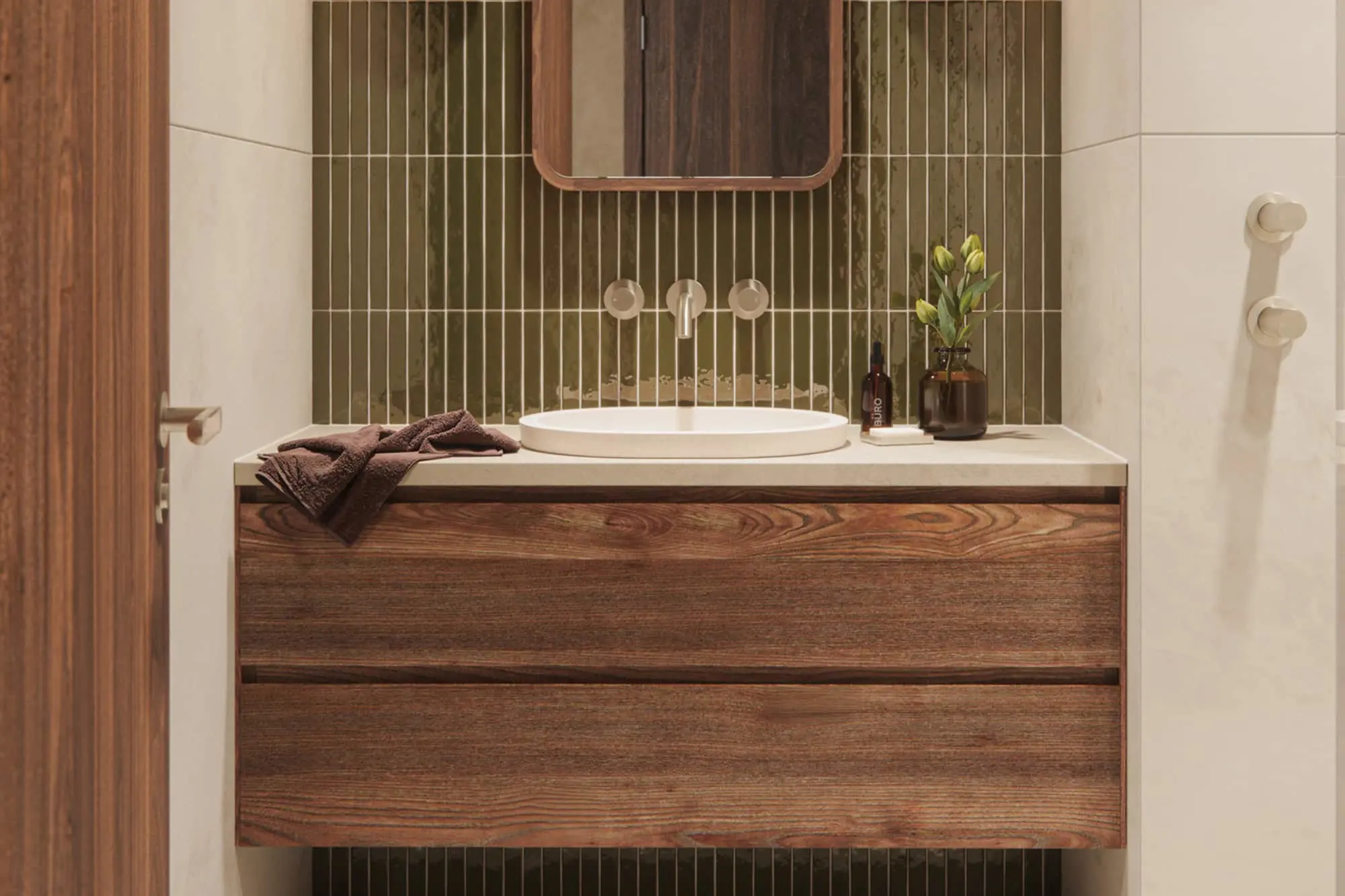 Bathroom vanity with a wood cabinet, white basin, green vertical tiles, wall-mounted tap, mirror, and dark bottle with plant.
