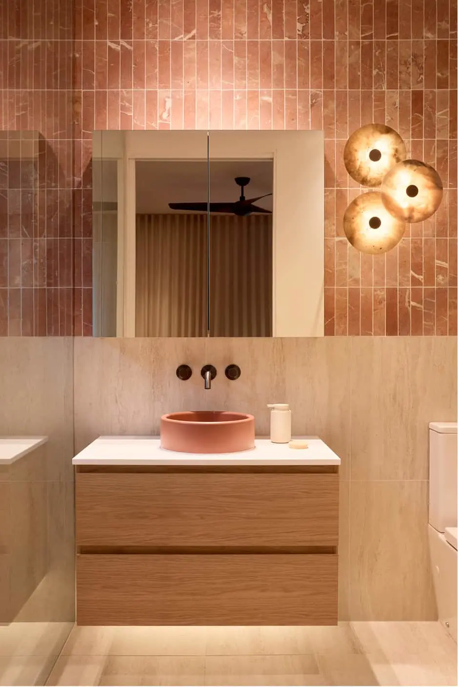 Bathroom with pink tiled walls, round coral sink on wood vanity, mirror, and three circular gold wall lights.
